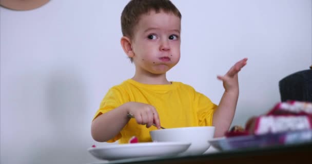 Μικρό χαριτωμένο παιδί κάθεται στο τραπέζι και τρώει το δικό του πλιγούρι βρώμης, και τρώει εξωτικά φρούτα, το μωρό τρώει πρόθυμα. Η έννοια της ευτυχης παιδικής ηλικίας. — Αρχείο Βίντεο
