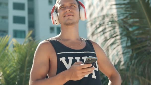 Joven guapo escuchando música desde su teléfono inteligente con auriculares blancos inalámbricos, bailando en la playa urbana en el fondo Blue Sky . — Vídeo de stock