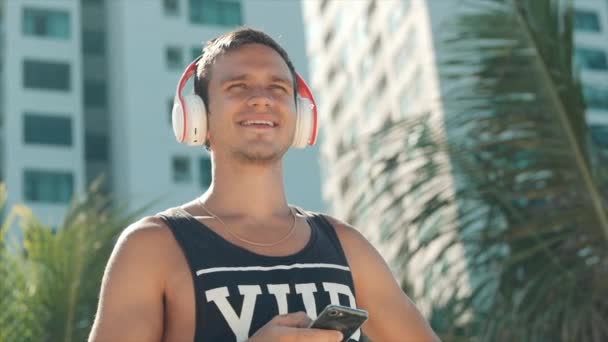 Joven guapo escuchando música desde su teléfono inteligente con auriculares blancos inalámbricos, bailando en la playa urbana en el fondo Blue Sky . — Vídeos de Stock