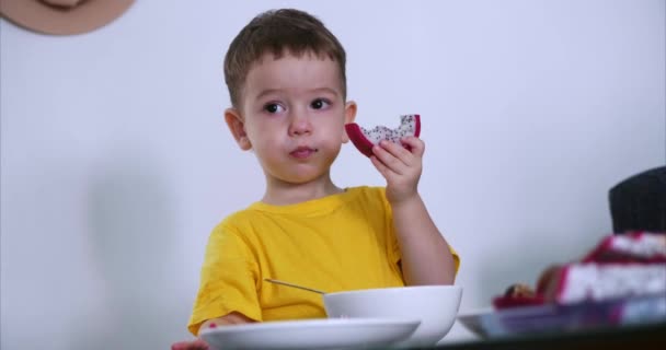 Маленький милый ребенок садится за стол и ест свою собственную овсянку, и ест экзотические фрукты, ребенок ест добровольно. Концепция счастливого детства . — стоковое видео