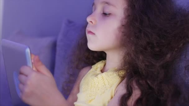 Foto nocturna Retrato de un niño lindo caucásico, niña pequeña de cerca de una cara de niño Viendo una tableta PC con un reflejo y fotos en sus ojos . — Vídeo de stock