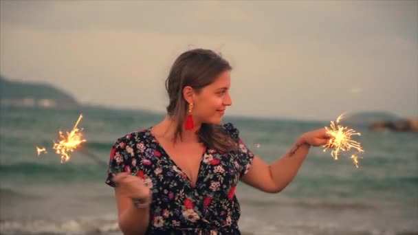 Joven mujer sonriente feliz, bailando con chispeante al atardecer en cámara lenta, con fuegos artificiales al atardecer en la playa . — Vídeos de Stock