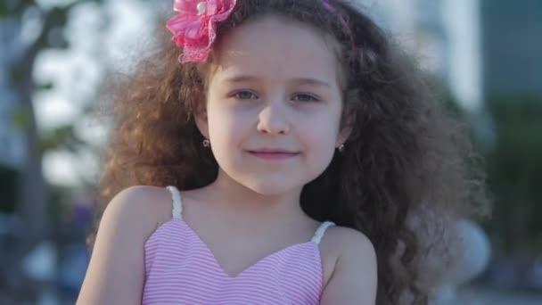 Conceito Infância feliz. Retrato bonito pequena criança caucasiana fêmea, criança ao ar livre em um vestido rosa com uma flor rosa em sua cabeça, olhando na câmera. Conceito Infância feliz . — Vídeo de Stock