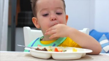 Sevimli küçük çocuk bir masada bir Bib oturan ve kendi püresi Eat, Bebek Isteyerek yiyor. Tatlı küçük bebek yemek onu yemek. Konsept mutlu Childhool.