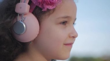 Kıvırcık saçlı bir sevimli çocuk portresi, pembe elbise içinde bir pembe çiçek Ile beyaz küçük kız saçlı, pembe kulaklıklar bebek müzik dinleme, kamera bakıyor, tatlı gülümseyen