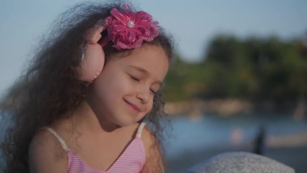 Portret van een schattig kind met krullend haar, mooie Kaukasische meisje zomer in een roze jurk met een roze bloem op haar hoofd in haar haar, baby in Pink koptelefoon luisteren naar muziek, glimlachend zoet. — Stockvideo