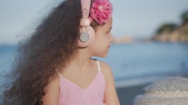 Kıvırcık saçlı bir sevimli çocuk portre, beatiful Kafkas küçük kız yaz pembe elbise bir pembe çiçek saç, Bebek pembe kulaklıklar içinde onun kafası Ile müzik dinleyin, tatlı gülümseyen.