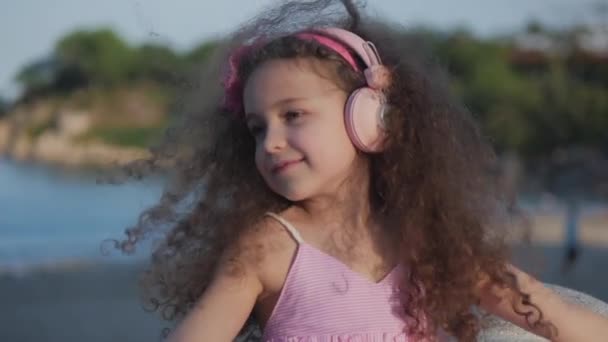 Ritratto di un bambino carino con i capelli ricci, Beatiful caucasico bambina estate in un abito rosa con un fiore rosa sulla testa tra i capelli, Bambino in cuffie rosa Ascoltare musica, Sorridente dolcemente . — Video Stock