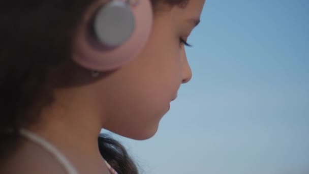 Retrato de uma criança bonito com cabelo encaracolado, menina branca em um vestido rosa com uma flor rosa em sua cabeça em seu cabelo, bebê em fones de ouvido rosa Ouvindo música, Olhando para a câmera, Sorrindo docemente — Vídeo de Stock