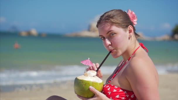 Nahaufnahme Porträt einer schönen, brünetten jungen Frau aus Europa oder eines fröhlichen Mädchens, das eine Kokosnuss in der Sonne im Wind an einem tropischen Strand trinkt. Konzept Meereswellen, die im Sommer am Meer entspannen. — Stockvideo