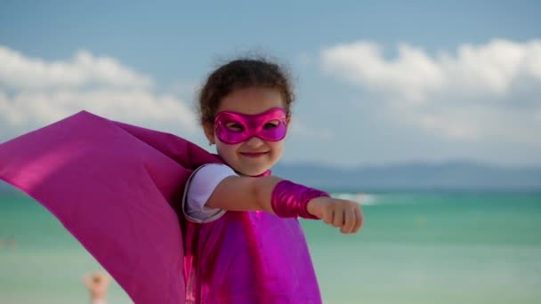 Mooi klein meisje in de superheld kostuum, gekleed in een roze mantel en het masker van de held. Speelt op de achtergrond zee en blauwe lucht en wolken, stuurt een vuist naar voren. Concept van een gelukkig — Stockvideo