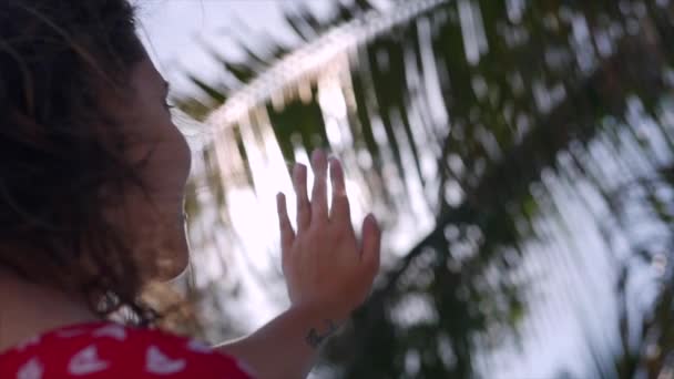 Close-Up Retrato de uma bela morena bonita europeia Jovem ou uma menina alegre, fecha a mão do sol que brilha através dos dedos e folhas de árvores, no vento em uma praia tropical . — Vídeo de Stock
