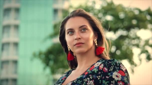 Fermer Portrait Une merveilleuse femme aux cheveux bruns aux yeux verts avec des cheveux fluides dans une robe d'été avec une image de fleurs rouges et blanches contre l'arbre vert et le bâtiment gris-vert. . Concentration douce — Video