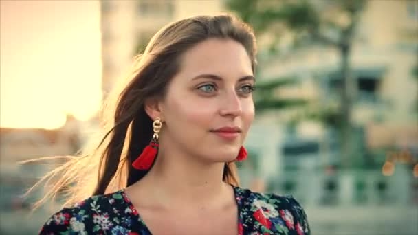 Close up Retrato uma mulher de cabelos castanhos maravilhosa com olhos verdes com cabelo fluindo em um vestido de verão com uma imagem de flores vermelhas e brancas contra árvore verde e edifício verde-cinza no Set Sun . — Vídeo de Stock