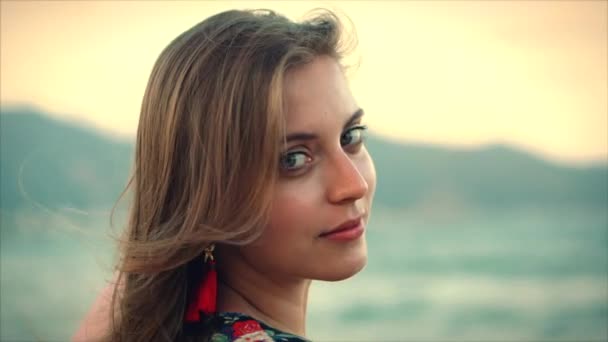 Close up Retrato uma bela mulher de cabelos castanhos com olhos verdes com cabelo fluindo em um vestido de verão com uma imagem de flores vermelhas e brancas girando ao redor com cachecol em uma praia em Set Sun . — Vídeo de Stock