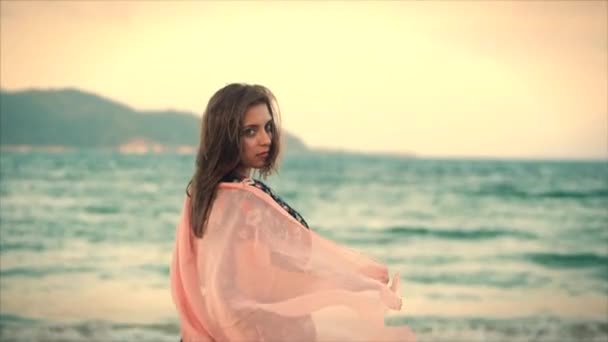 Close-up portret een mooie bruinharige vrouw met groene ogen met vloeiende haren in een zomerjurk met een foto van rode en witte bloemen ronddraaien met sjaal op een strand in de set zon. zachte focus — Stockvideo