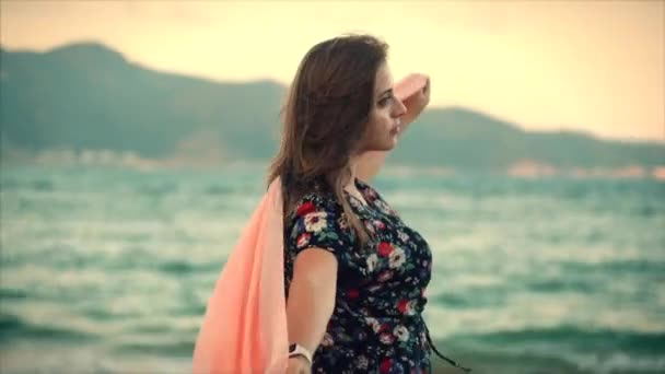 De cerca Retrato de una hermosa mujer de cabello castaño con los ojos verdes con el pelo que fluye en un vestido de verano con una imagen de flores rojas y blancas girando alrededor con bufanda en una playa en Set Sun.Soft foco — Vídeo de stock