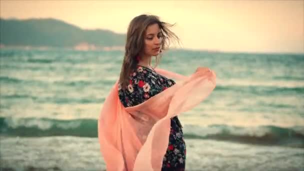 Close-up portret een mooie bruinharige vrouw met groene ogen met vloeiende haren in een zomerjurk met een foto van rode en witte bloemen ronddraaien met sjaal op een strand in de set zon. zachte focus — Stockvideo