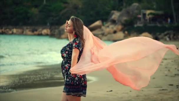 Close-up portret een mooie bruinharige vrouw met groene ogen met vloeiende haren in een zomerjurk met een foto van rode en witte bloemen ronddraaien met sjaal op een strand in de set zon. zachte focus — Stockvideo