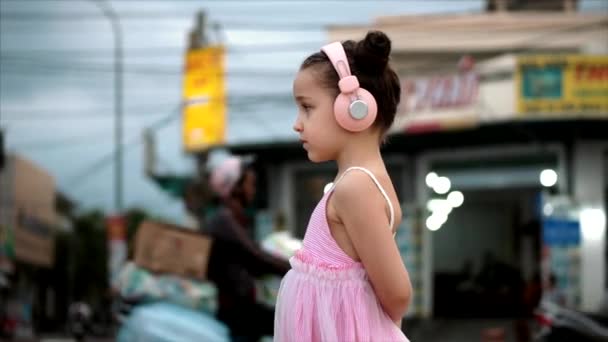 Portrait d'un enfant mignon debout près de la route, une merveilleuse petite fille mignonne en robe rose et écouteurs roses regardant loin, sur fond de voitures et de motos passantes . — Video