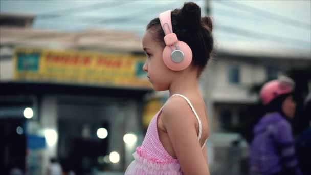 Retrato de uma criança bonita em pé na estrada, uma menina maravilhosa bonito em um vestido rosa e fones de ouvido rosa olhando para fora, contra o fundo de passar carros e motocicletas . — Vídeo de Stock