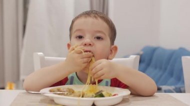 Küçük çocuk bir masada bir Bib oturan ve kendi spagetti Eat, sevimli bebek Isteyerek yiyor. Tatlı küçük bebek yemek onu yemek. 4k