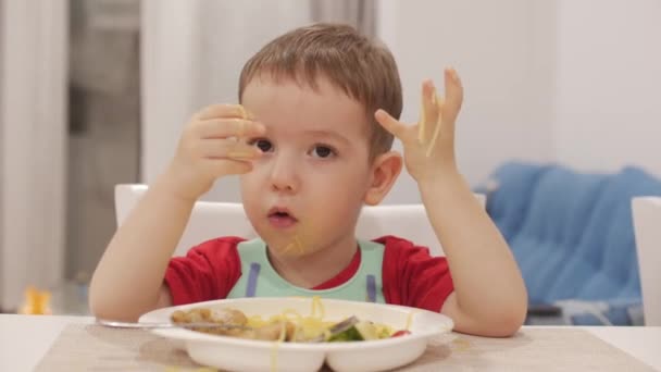 Criança pequena está sentada em uma mesa em um babete e comer seu próprio espaguete, o bebê bonito come voluntariamente. Bebé bonito comendo seu jantar. 4K — Vídeo de Stock