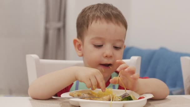 Små barn sitter vid ett bord i en haklapp och äta sin egen spaghetti, den söta baby Eats villigt. Söt liten bebis äta hennes middag. 4k — Stockvideo