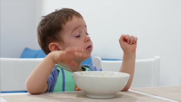 Klein schattig kind zit aan een tafel en eet zijn eigen havermout, de baby eet gewillige. Concept gelukkige jeugd. — Stockvideo