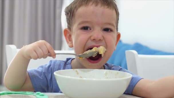 Criança pequena bonito está sentado em uma mesa e comer sua própria aveia, o bebê come de bom grado. Conceito Infância feliz . — Vídeo de Stock