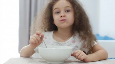 Small Cute Girl with Curly Hair is Sitting at a Table and eat his own oatmeal, the Child Eats Willingly. Concept Happy Childhood.