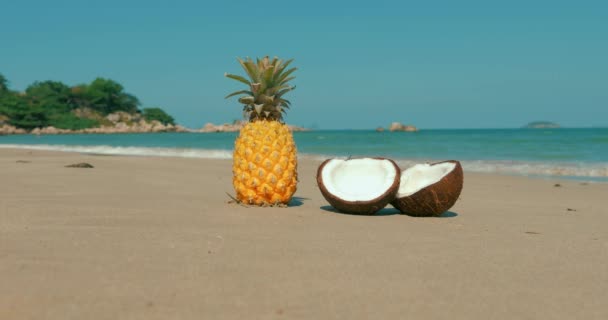 An einem tropischen Strand in Nahaufnahme unter der heißen Sommersonne entlang der tropischen exotischen Küste, auf dem Sand stehend Ananas und in der Hälfte Kokosnuss auf dem Meeresgrund gespalten. Konzept aktuell, Sommer, Urlaub. — Stockvideo