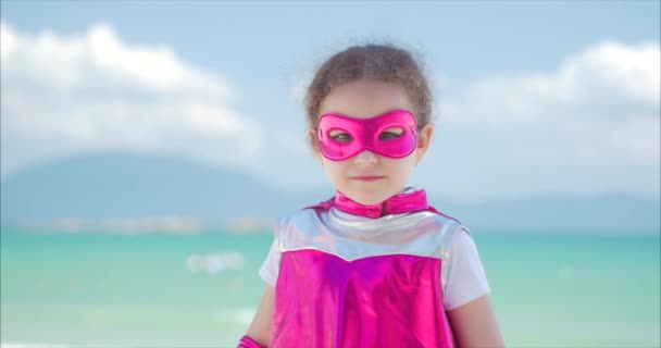 Mooi klein meisje in de superheld kostuum, gekleed in een roze mantel en het masker van de held. Speelt op de achtergrond zee en blauwe lucht en wolken, stuurt een vuist naar voren. Concept van een gelukkig — Stockvideo