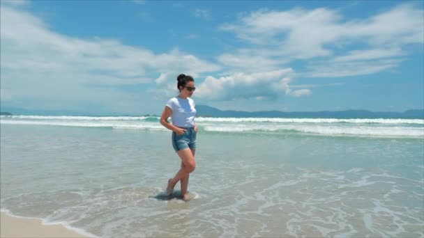 Europese mooie brunette in zwarte glazen, wit-shirt en blauwe broek gaat onder de hete zomer zon langs de tropische exotische kust van Vietnam, zorgeloos meisje lopen op het zand recht op de Oceaan — Stockvideo