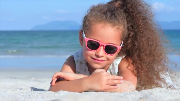 ピンクのメガネをかけた美しい少女のクローズアップポートレート、カメラを見てかわいい笑顔、海辺の砂の上に横たわって、手の砂を注ぎます。コンセプト:子供、子供時代、夏、子供、子供. — ストック動画