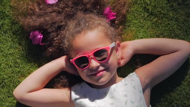 Portrait d'un enfant mignon, une merveilleuse petite belle fille dans une robe blanche et des lunettes roses, allongé sur l'herbe regardant l'appareil photo et souriant doucement. Concept de l'enfance heureuse . — Video