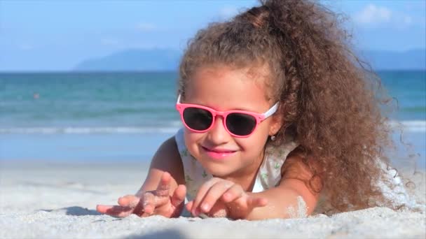 Närbild porträtt av en vacker liten flicka i rosa Glasögon, söt leende tittar på kameran, liggande på sanden vid havet, häller sand av hand. Koncept: barn, Childhood, sommar, barn, Kid. — Stockvideo