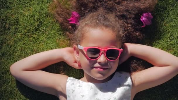 Retrato de un niño lindo, una niña hermosa maravillosa en un vestido blanco y gafas de color rosa, acostado en la hierba mirando a la cámara y sonriendo dulcemente. Concepto de la Infancia Feliz . — Vídeos de Stock