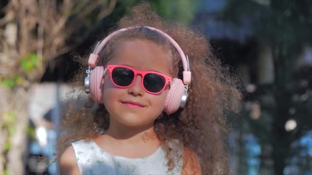 Porträt eines süßen Kindes, eines wunderbaren kleinen schönen Mädchens in einem weißen Kleid mit rosa Brille und rosa Kopfhörern, das in die Kamera schaut und Musik hört. Konzept glückliche Kindheit. — Stockvideo