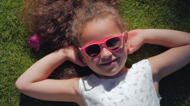 Portrait d'un enfant mignon, une merveilleuse petite belle fille dans une robe blanche et des lunettes roses, allongé sur l'herbe regardant l'appareil photo et souriant doucement. Concept de l'enfance heureuse . — Video