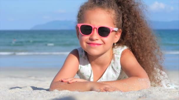 Nahaufnahme Porträt eines schönen kleinen Mädchens mit rosa Brille, niedlich lächelnd in die Kamera blickend, auf dem Sand am Meer liegend, schüttet Sand von Hand. Konzept: Kinder, Kindheit, Sommer, Kind, Kind. — Stockvideo