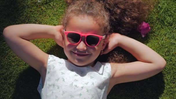Portrét roztomilýho dítěte, nádherné malé krásné dívky v bílých šatech a růžových brýlích, ležící na trávě, dívajíce se na kameru a s úsměvem sladce. Koncept šťastného dětství. — Stock video