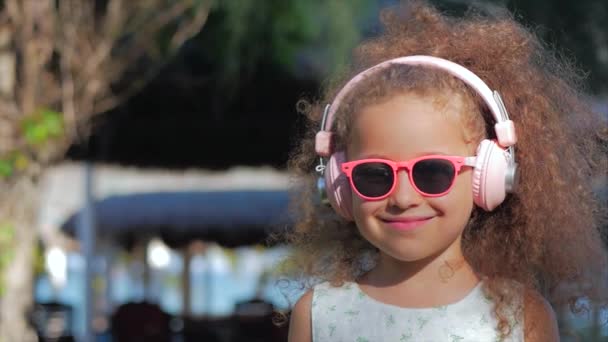 Ritratto di un bambino carino, una meravigliosa bambina in un abito bianco con occhiali rosa e cuffie rosa, Guardando la fotocamera, Ascoltando la musica. Concetto Infanzia felice . — Video Stock