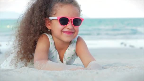 Close-up portret van een mooi klein meisje in roze bril, schattig glimlachend kijkend naar de camera, liggend op het zand aan zee, giet zand van de hand. Concept: kinderen, jeugd, zomer, kind, Kid. — Stockvideo