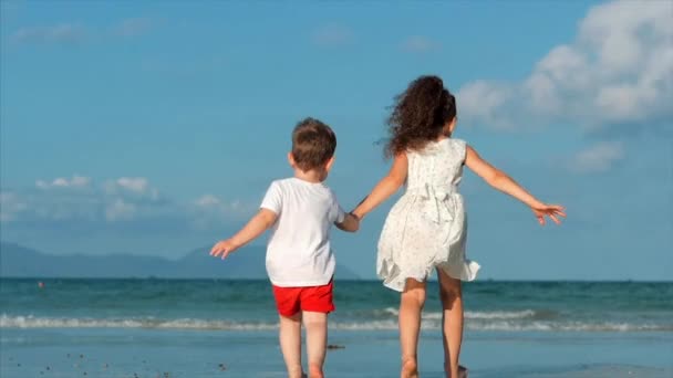Milé děti, děvče a chlapec držící ruce jeden druhého běží podél tropického pobřeží k oceánu. Koncept: děti, šťastné dětství, léto, Baby, dovolená. — Stock video