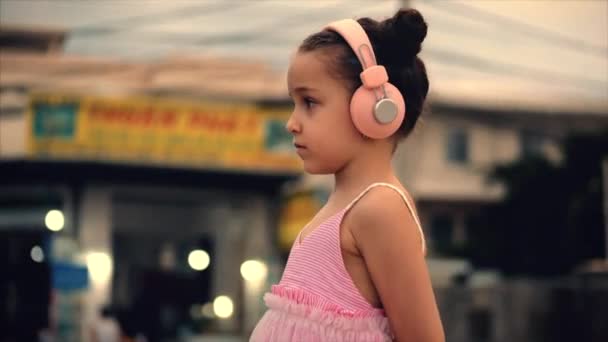 Retrato de uma criança bonita em pé na estrada, uma menina maravilhosa bonito em um vestido rosa e fones de ouvido rosa olhando para fora, contra o fundo de passar carros e motocicletas . — Vídeo de Stock