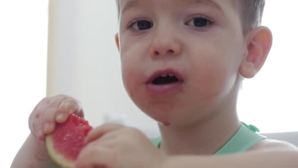 Ein kleines Kind sitzt in einem Lätzchen an einem Tisch und isst seine eigene Wassermelone, das süße Baby isst bereitwillig. niedliches kleines Baby, das ihr Abendessen isst. 4k — Stockvideo