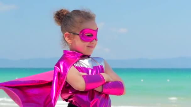 Vacker liten flicka i superhjälte kostym, klädd i en röd kappa och masken av hjälten. Spelar på bakgrunden havet och blå himmel och moln, skickar en knytnäve framåt. Begreppet Happy chilhood. — Stockvideo