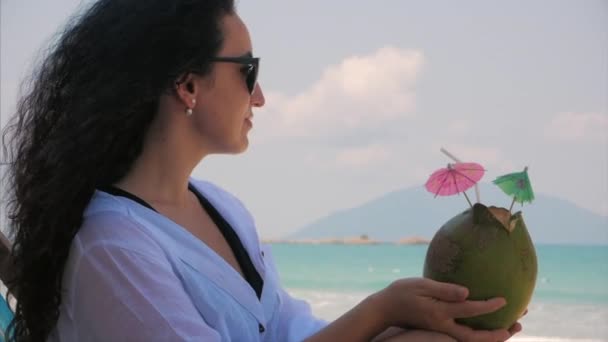 一个欧洲美丽的可爱布鲁内特年轻女子或一个快乐的女孩的肖像，在阳光下喝椰子，在热带海滩的风。概念夏季， 假期， 旅行. — 图库视频影像