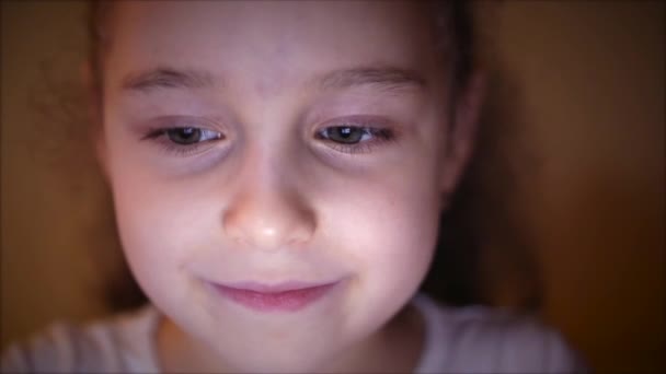 Tournage nocturne d'une jolie petite fille caucasienne Gros plan d'un visage d'enfant regardant une tablette PC avec un reflet de lutte et des photos dans ses yeux — Video